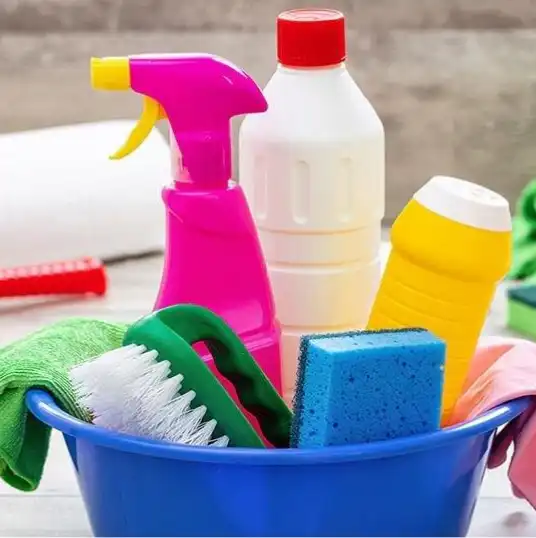 Cleaning-Products-2 Blue bucket filled with cleaning supplies: pink spray bottle, white bottle with red cap, yellow bottle, sponge, and brush with gloves nearby.